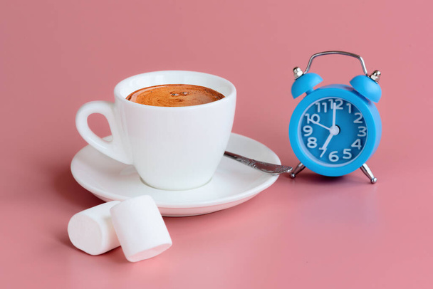 Morning cup of coffee, two marshmallows and blue alarm clock on pink background. 7 o'clock on clock face. Start of the day concept. Copy space - Photo, Image