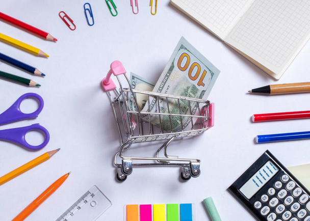 Top view photo of a small shopping cart with dollars in the center and colorful office supplies, isolated on white with copy space. Back to school concept. School shopping concept - Foto, afbeelding