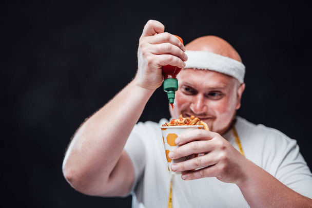 Homme en surpoids avec un bandage blanc sur la tête et en vêtements de sport, manger des frites - Photo, image