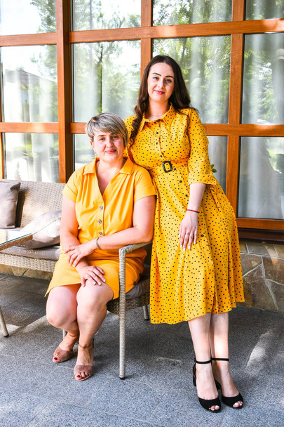 Mother and daughter in yellow dresses sitting outdoors. Family portrait. Looking at camera. Two women smiling. Pretty Brunette with long hair and Her Mom, Mothers day - Foto, Imagen