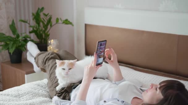 jeune femme utilise smartphone et commande de la nourriture en ligne en utilisant Internet tout en étant allongé à la maison sur le lit avec chat pendant l'auto-isolement - Séquence, vidéo
