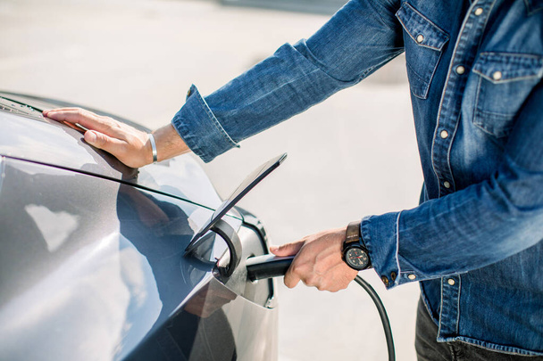 Close up cropped image of hands of man in jeans shirt and his electric car at charging station with the power cable supply plugged in. Electric cars, future car concept - Foto, Imagen