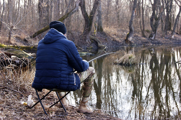 Wędka w rękach rybaka na jeziorze. Młody facet siedzi na krześle z wirującym prętem na tle wody.. - Zdjęcie, obraz