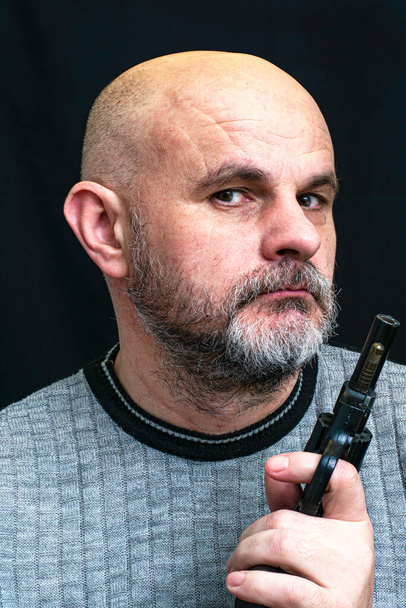 calvo barbudo hombre de mediana edad con una pistola en la mano sobre el fondo oscuro
 - Foto, Imagen