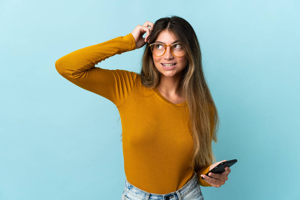 Mujer joven caucásica usando teléfono móvil aislado sobre fondo azul que tiene dudas y con expresión de cara confusa
 - Foto, imagen