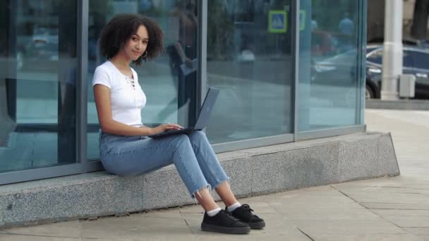 Приятные молодые положительные кудрявые волосы африканской этнической женщины наслаждаясь работой на ноутбуке улыбаясь показывая хорошо знак большой палец вверх, дает отличную оценку высокого качества сидя на отражательное окно на открытом воздухе - Кадры, видео