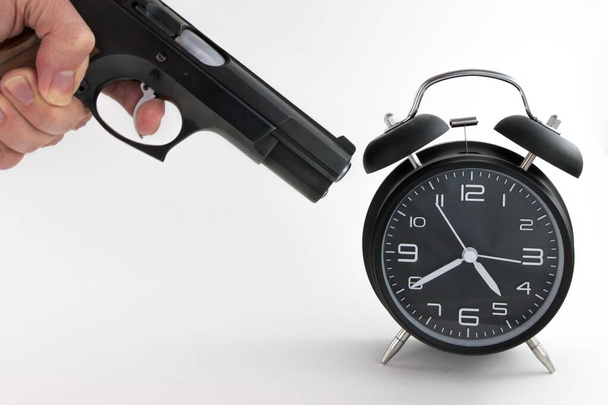 A close up shot of a person trying to destroy an alarm clock by a gun- working responsibilities and deadline concept - Photo, Image