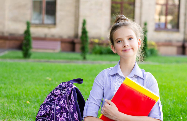 Vissza a suliba. Oktatási koncepció. Aranyos mosolygós iskoláslány ül a füvön az iskola mellett. Boldog kislány gyermek kezében a könyveket hátizsák - Fotó, kép