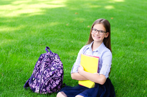 Back to school. Education concept. Cute smiling schoolgirl in glasses sitting on grass. Happy little girl child holding the books - Foto, afbeelding