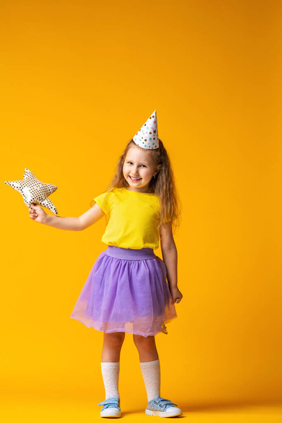 happy Birthday! Studio portrait of a little girl in a festive hat on her birthday on a yellow background. the child smiles and holds a textile star in his hands. Waiting for a gift. - Photo, image
