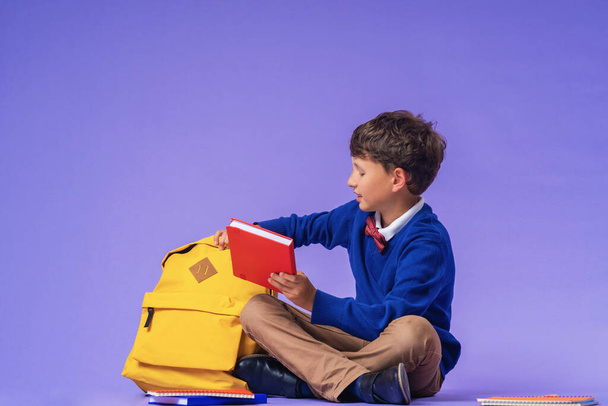 Cute serious schoolboy puts office supplies in a backpack on a purple background. Preparation for school. Back to school. Self-Assembly of a school backpack. - Фото, зображення
