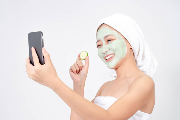Aziatische vrouw is gezichtsmasker. Ze maakt een foto met een verse komkommer op een witte achtergrond. - Foto, afbeelding