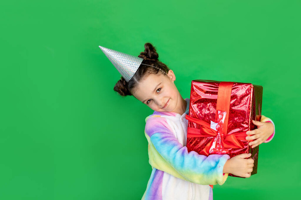 una graziosa bambina su uno sfondo verde isolato in un abito luminoso e con un cappello in possesso di un regalo. Spazio per il testo. Nuovo anno e concetto di Natale - Foto, immagini