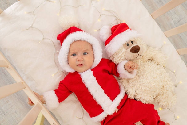 Boże Narodzenie zdjęcie dziecka w stroju Mikołaja leżącego w łóżeczku w domu z zabawką w kapeluszu Świętego Mikołaja, widok z góry, Szczęśliwego Nowego Roku. - Zdjęcie, obraz