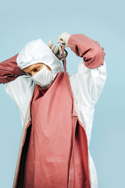 Fully geared up doctor in a white costume covering all skin, wearing mask, gloves and glasses. He's putting on red surgeon apron tying laces behind his neck. - 写真・画像