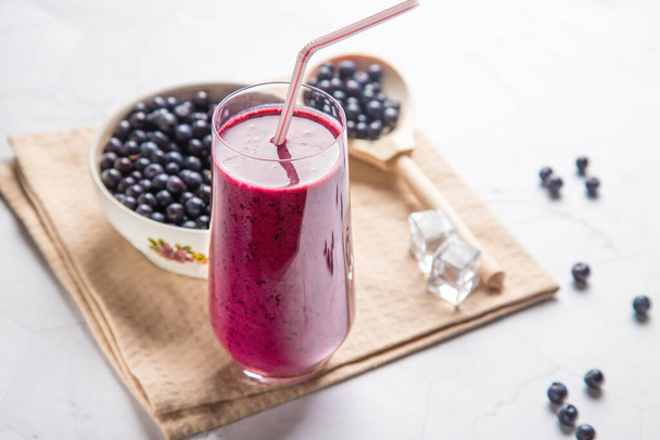 Diet drink blueberry smoothie in a glass glass with a tube, white plate with berries on a light background, with a beige napkin, in a wooden spoon-blueberries - Photo, Image