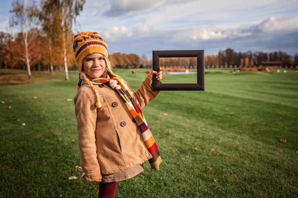 Podzimní zralost je mistrovské dílo. Roztomilá dívka v jasném šátku a klobouku s dřevěným rámem zvýrazňující krásu přírody, krásné zlaté podzimní krajiny, ekologie a životního prostředí - Fotografie, Obrázek