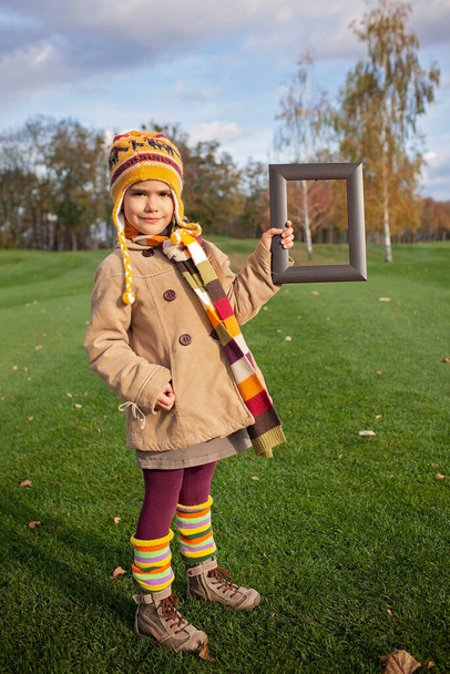 O outono maduro é obra-prima. Menina bonito em cachecol brilhante e chapéu segurando uma moldura de madeira para acentuar a beleza da natureza, bela paisagem dourada outono, ecologia e conceito de meio ambiente
 - Foto, Imagem