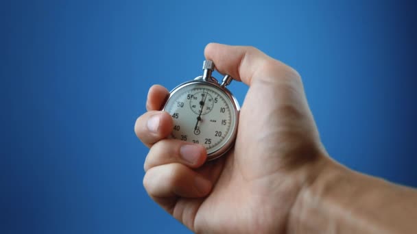 Reloj de mano masculino con cronómetro analógico sobre fondo de color azul. El tiempo comienza con el viejo cronómetro hombre presiona el botón de inicio en el concepto de deporte. Concepto de gestión del tiempo. - Imágenes, Vídeo