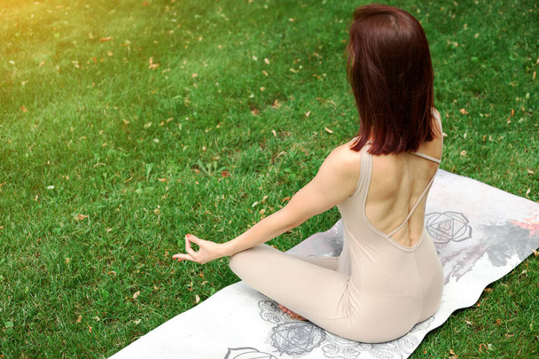 Healthy, yoga and meditation concept. Back view European beautiful woman sitting in lotus position on mat wearing sportswear and relaxing in the city park at summer morning day. Empty copy space. - Foto, imagen