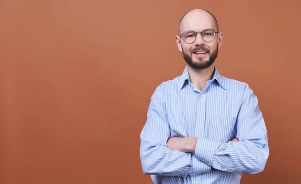 A handsome young man, a businessman, bald, with a beard, in round glasses and a striped shirt, crossed his arms on his chest and looks straight. Close-up of the face of happy successful business man. - Foto, immagini