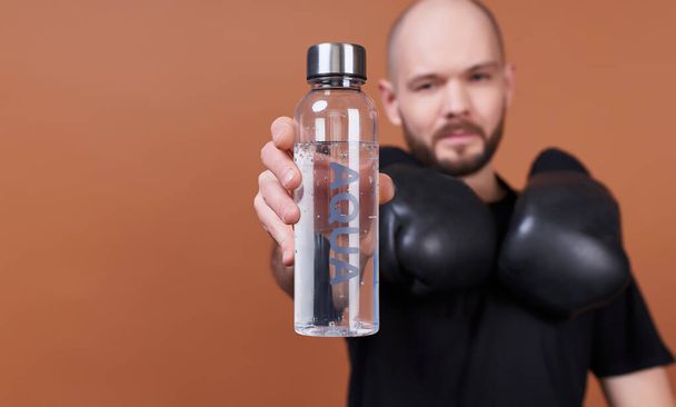 Portrait of a charming smiling guy with a beard, in a black T-shirt, he after training on boxing, holding a water bottle with the inscription aqua. The concept of sports and water balance, lifestyle. - Фото, изображение
