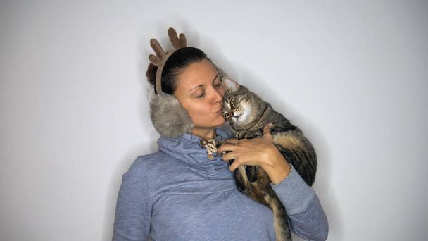 Una mujer con orejeras de reno besa a su gato rayado que sostiene en sus brazos. - Foto, imagen