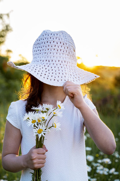 a beautiful little girl with daisies covers her face with a hat - Foto, Bild