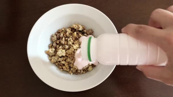 Dry muesli in a white plate on a brown wooden table. Kefir is poured into muesli. A woman's hand pours dry muesli with yogurt and mixes them with a silver spoon - Video, Çekim