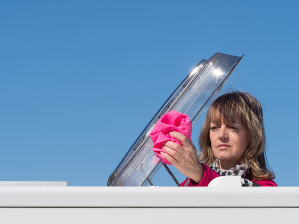 Una signora proprietario camper pulisce il portello lucernario del suo veicolo con un panno rosa.Testa e spalle sono visibili come lei è salita attraverso il lucernario tetto.Copia spazio.Immagine - Foto, immagini