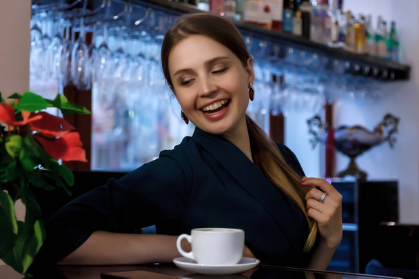 Girl of Slavic appearance in bar, cafe or restaurant with white Cup. Female shows emotion. Young charming woman is sitting at bar and drinking coffee or tea in her free evening time - Photo, image