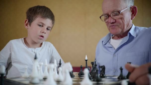 board games, an elderly man in glasses for vision and a happy cute male child have fun together playing chess together - Video, Çekim