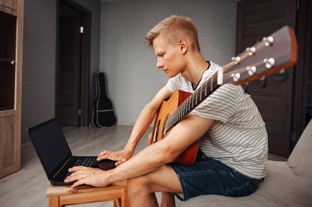 Focused blond man learning to play guitar online using laptop while sitting at home, distance learning - Fotografie, Obrázek