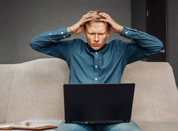 Upset young man clutching his head due to hard work at the computer while sitting at home on the couch, working remotely at home - Φωτογραφία, εικόνα