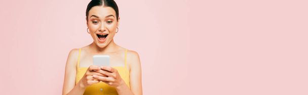 shocked brunette young woman using smartphone isolated on pink, panoramic shot - 写真・画像
