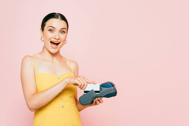 excited brunette young woman using credit card and payment terminal isolated on pink - Fotoğraf, Görsel