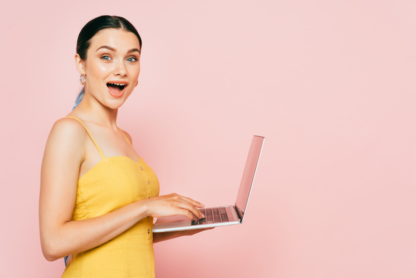 Seitenansicht einer schockierten brünetten jungen Frau mit Laptop auf rosa - Foto, Bild