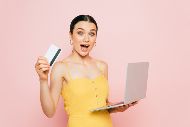 excitada morena joven sosteniendo tarjeta de crédito y portátil en rosa - Foto, Imagen