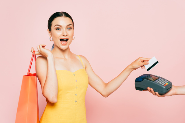 σοκαρισμένη μελαχρινή νεαρή γυναίκα με τσάντα ψώνια που πληρώνει με πιστωτική κάρτα σε ροζ - Φωτογραφία, εικόνα