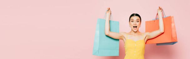 vzrušená brunetka mladá žena s nákupními taškami ve zvednutých rukou na růžové, panoramatický výstřel - Fotografie, Obrázek