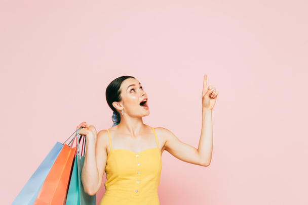 impactado morena joven con bolsas de compras apuntando hacia arriba en rosa - Foto, Imagen