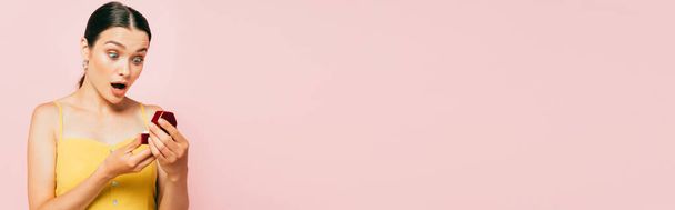 chocado morena jovem segurando caixa com anel de noivado isolado em rosa, tiro panorâmico
 - Foto, Imagem