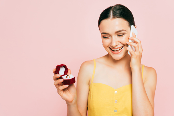 brunette young woman holding box with engagement ring while talking on smartphone isolated on pink - Foto, Imagen