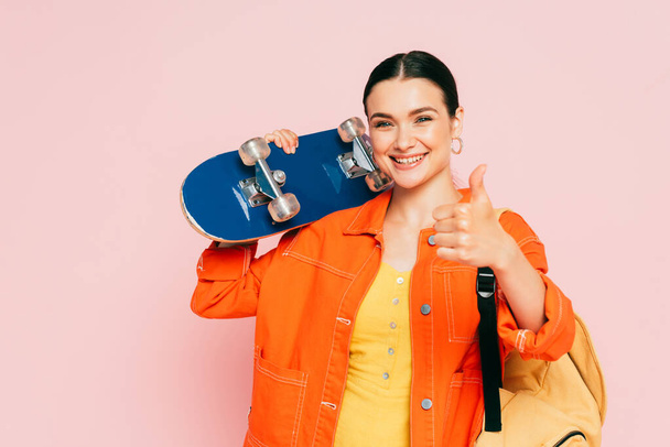 brunette young woman in colorful outfit with skateboard showing thumb up isolated on pink - Foto, Imagen