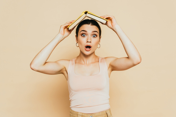 shocked brunette young woman with book on head on beige - Foto, Imagen