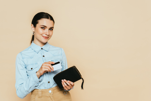 brünette Frau im Jeanshemd mit Geldbörse und Kreditkarte isoliert auf beige - Foto, Bild
