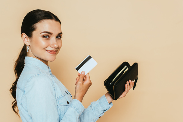 side view of brunette woman in denim shirt holding wallet and credit card isolated on beige - Foto, Imagem