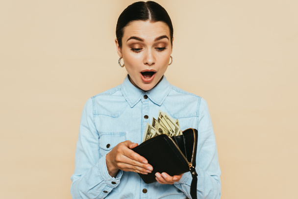 shocked brunette woman in denim shirt holding wallet with dollars isolated on beige - Foto, Imagem