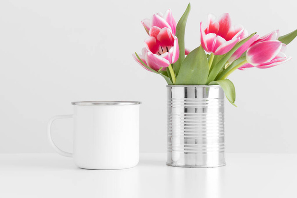 Enamel mug mockup with pink tulips in a pot on a white table. - Photo, Image