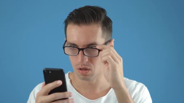 Güneş Gözlüklü Şok Adam Smartphone 'da tepki veriyor. Nahoş bir şekilde şaşırmış ve korkmuştu. Erkek, mavi arka planda, akıllı telefonunda gördüklerine şaşırır.. - Video, Çekim
